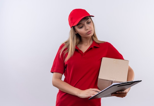 Denkende jonge levering meisje dragen rode uniform en GLB bedrijf doos en kijken naar klembord in haar hand geïsoleerd op wit