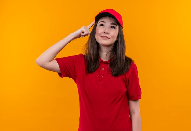 Denkende jonge leveringsvrouw die rode t-shirt in rode GLB draagt op geïsoleerde oranje muur