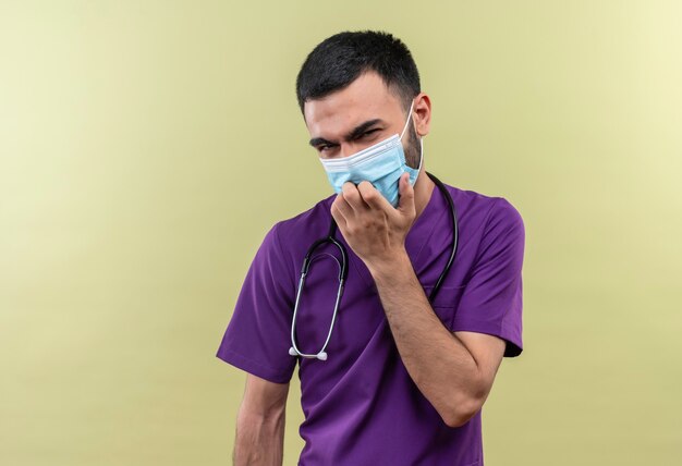 Denkende jonge mannelijke arts die paarse chirurgenkleding en een stethoscoop medisch masker draagt, legde zijn hand op de kin op geïsoleerde groene muur