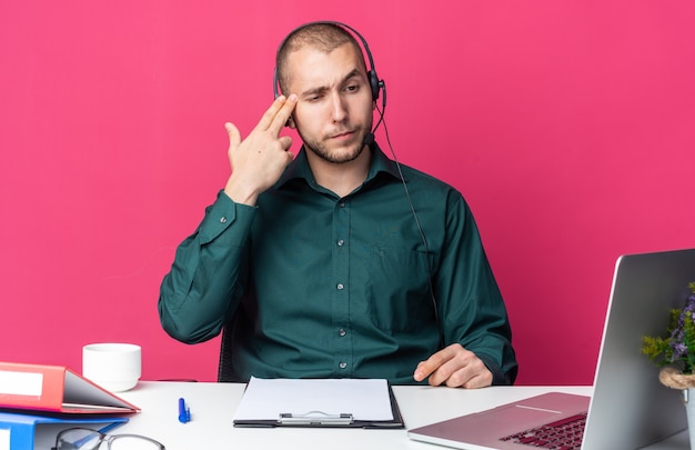 Denkende jonge mannelijke callcentermedewerker met een headset die aan een bureau zit met kantoorhulpmiddelen en naar een laptop kijkt die zelfmoordgebaar met een pistoolgebaar toont