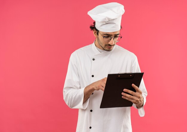 Denkende jonge mannelijke kok die eenvormige chef-kok en glazen dragen die en klembord bekijken dat op roze muur wordt geïsoleerd