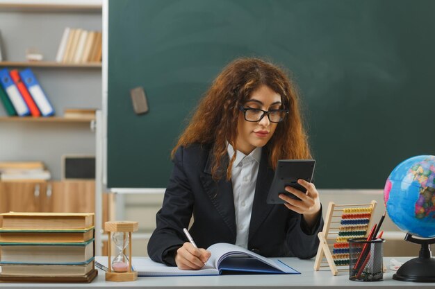 denkende jonge vrouwelijke leraar die een bril draagt en naar een rekenmachine kijkt die aan een bureau zit met schoolhulpmiddelen in de klas