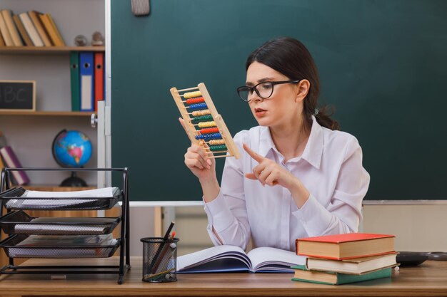 denkende jonge vrouwelijke leraar met een bril die een telraam vasthoudt en bekijkt dat aan een bureau zit met schoolhulpmiddelen in de klas