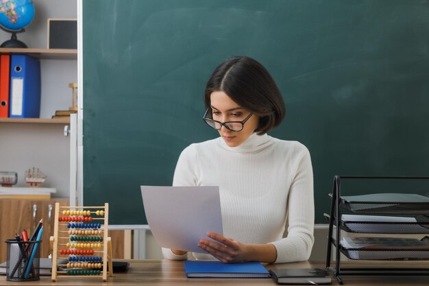 denkende jonge vrouwelijke leraar met een bril die papier vasthoudt en bekijkt terwijl hij aan het bureau zit met schoolhulpmiddelen in de klas