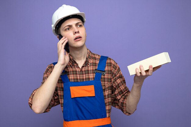 Denkende kijkende jonge mannelijke bouwer die een uniforme baksteen draagt, spreekt op de telefoon