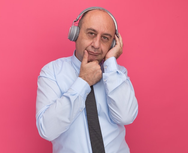 Denkende man van middelbare leeftijd met een wit t-shirt met stropdas en koptelefoon greep kin geïsoleerd op roze muur