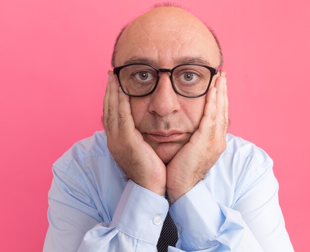 Denkende man van middelbare leeftijd met wit t-shirt met stropdas en bril handen op wangen geïsoleerd op roze muur