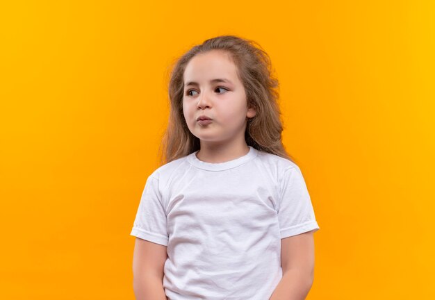 Denkende weinig schoolmeisje die witte t-shirt dragen die kant op geïsoleerde oranje muur bekijken