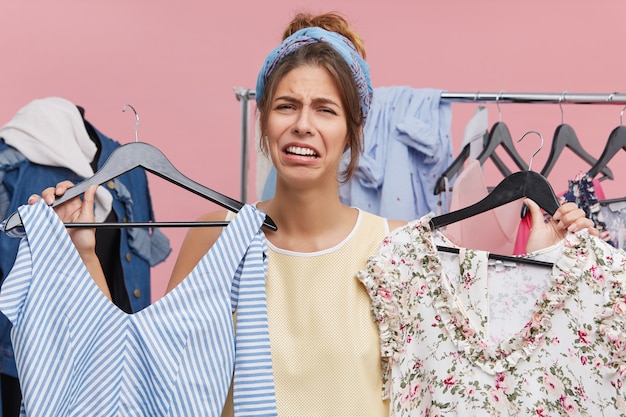 Gratis foto depressief droevig wijfje dat zich bij garderobe bevindt die twee hangers houdt met kledingstukken die beklemtoond voelen