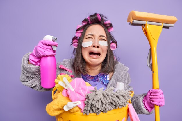 Depressieve Aziatische huisvrouw huilt uit wanhoop drukt negatieve emoties uit past patches onder de ogen toe om fijne lijntjes te verminderen draagt haarrollers poseert in de buurt van wasmand geïsoleerd over paarse muur