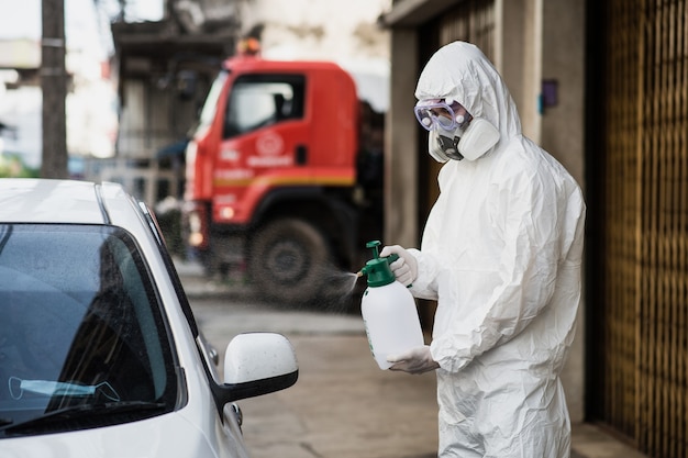 Desinfectie specialist man met persoonlijke beschermingsmiddelen (pbm) pak, handschoenen, masker en doorzichtige bril, schoonmaak auto met fles onder druk staand desinfectiemiddel om covid-19 te verwijderen