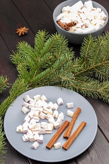 Dessert marshmallows en kaneelstokjes in keramische plaat en kom. pijnboom takken. donkere houten achtergrond.