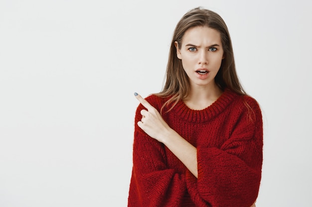 Die die dronken man uitnodigde om te feesten. Studio shot van ontevreden boze jonge vrouw in rode losse trui, fronsend en ruzie met vriend, wijzend op linkerbovenhoek