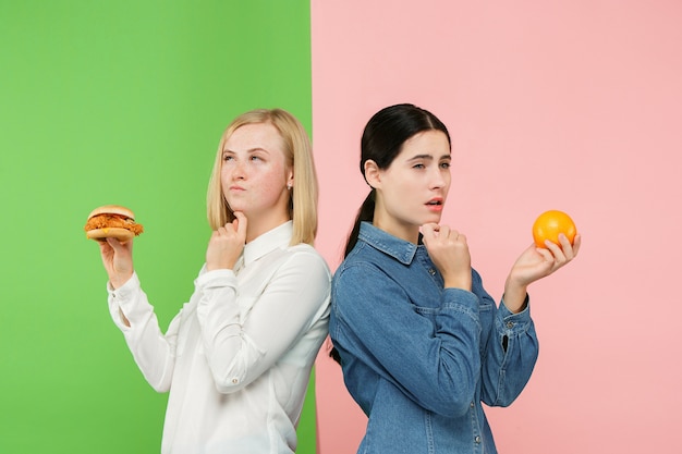 Dieet concept. Gezond, nuttig voedsel. Mooie jonge vrouwen kiezen tussen fruit en ongezond fastfood in de studio. Menselijke emoties en vergelijkingsconcepten