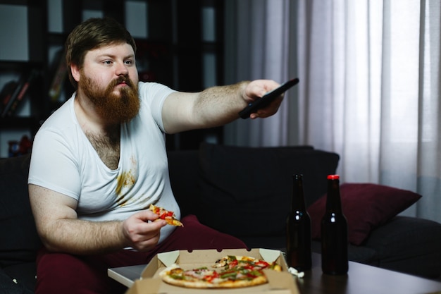 Dikke man eet pizza zittend op de bank en schakelt van kanaal op de tv