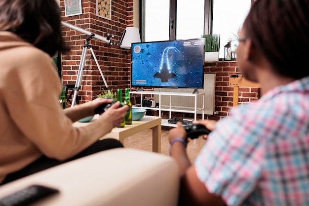 Diverse goede vrienden in de woonkamer zittend op de bank tijdens het spelen van space sim-videogame op een game-apparaat. Mensen die thuis zitten terwijl ze genieten van leuke vrijetijdsactiviteiten met een entertainmentconsole-apparaat.