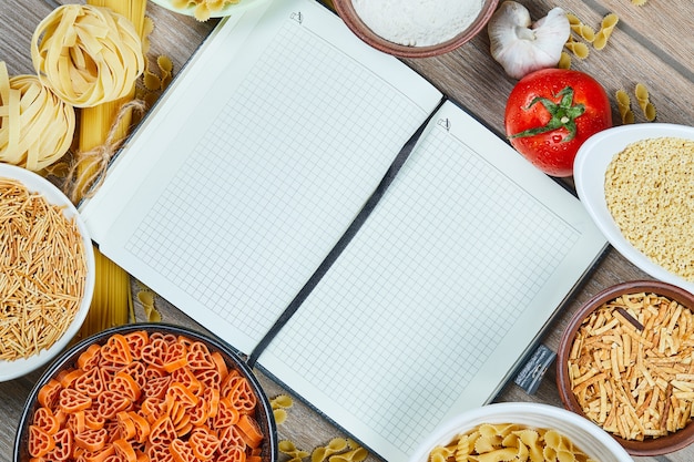 Diverse ongekookte deegwaren met notitieboekje en groenten op een houten lijst.