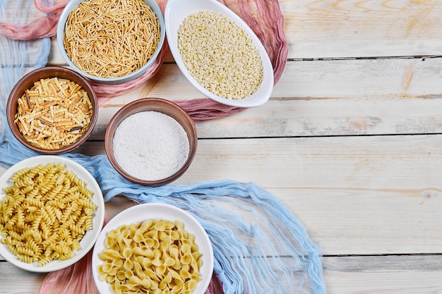 Diverse ongekookte pasta en meelkommen op houten tafel met blauw en roze tafelkleed.