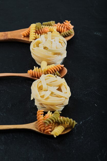 Diverse ongekookte pasta op houten lepels over donkere tafel.