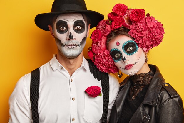 Dode vrouw en man dragen schedelmake-up, geschilderd voor Halloween, kijken verrassend naar de camera, gekleed in zwart-witte outfit voor Allerheiligen, geïsoleerd op gele achtergrond.