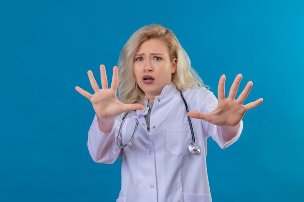 Doen schrikken jonge arts die stethoscoop in medische toga draagt die geen gebaar op blauwe muur toont