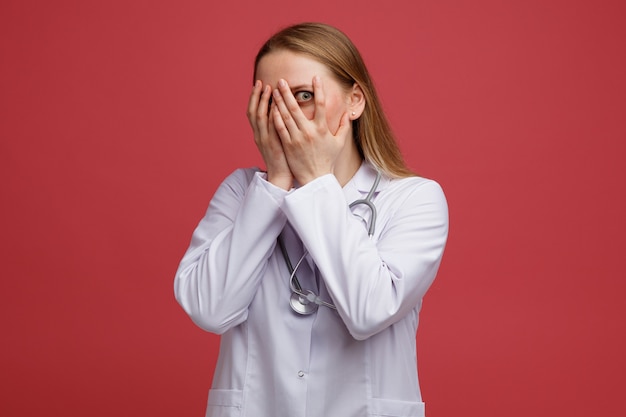 Doen schrikken jonge blonde vrouwelijke arts die medische mantel en een stethoscoop om de nek draagt die betrekking heeft op gezicht met handen die naar de camera tussen de vingers gluren