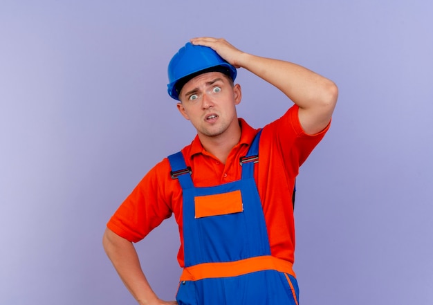 Doen schrikken jonge mannelijke bouwer die uniform en veiligheidshelm draagt die hand op hoofd op paars zetten