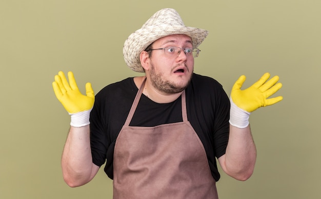 Gratis foto doen schrikken jonge mannelijke tuinman die het tuinieren hoed en handschoenen draagt die handen uitspreiden die op olijfgroene muur worden geïsoleerd