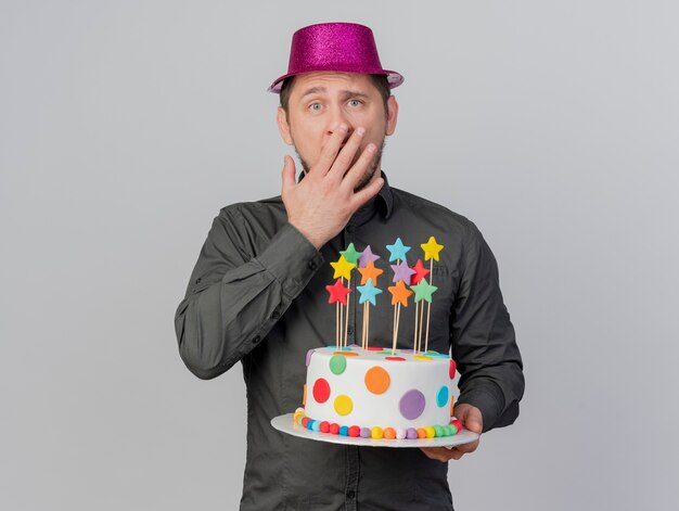 Doen schrikken jonge partij kerel die roze hoed draagt die cake behandelde mond met hand houdt die op witte achtergrond wordt geïsoleerd