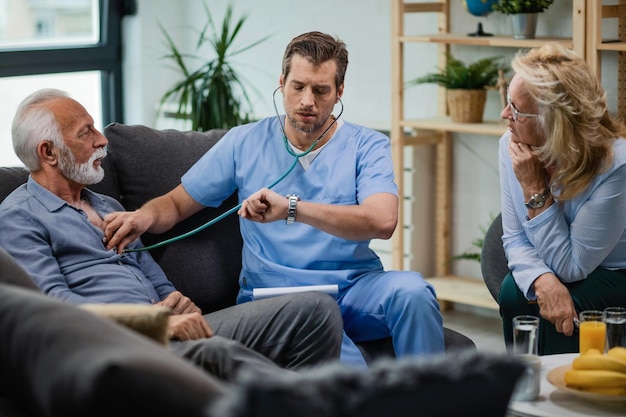 Dokter bezoekt senior koppel thuis en controleert zijn polshorloge terwijl hij luistert naar de hartslag van een man