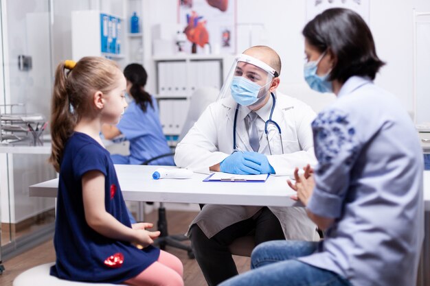 Dokter die gezichtsmasker draagt tijdens wereldwijde pandemie die tijdens overleg met kind praat. Gezondheid kinderarts specialist die gezondheidszorg consultaties behandeling in beschermende uitrusting.