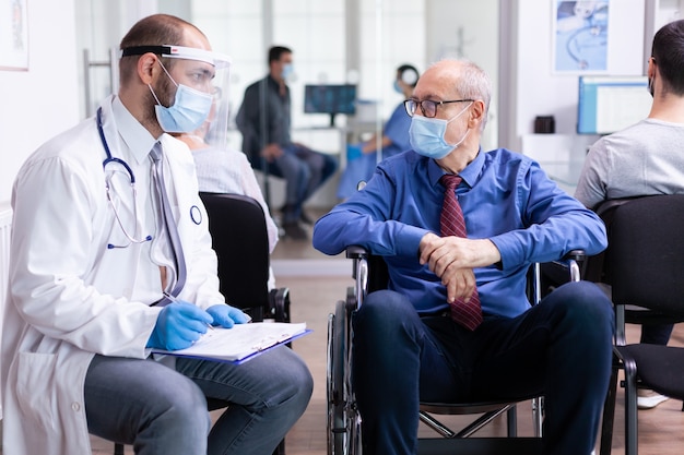 Dokter met gezichtsmasker en stethoscoop raadplegen gehandicapte senior man in wachtruimte van ziekenhuis