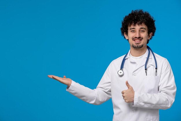 Doktersdag gekrulde knappe schattige kerel in medisch uniform glimlachend en zwaaiend met handen