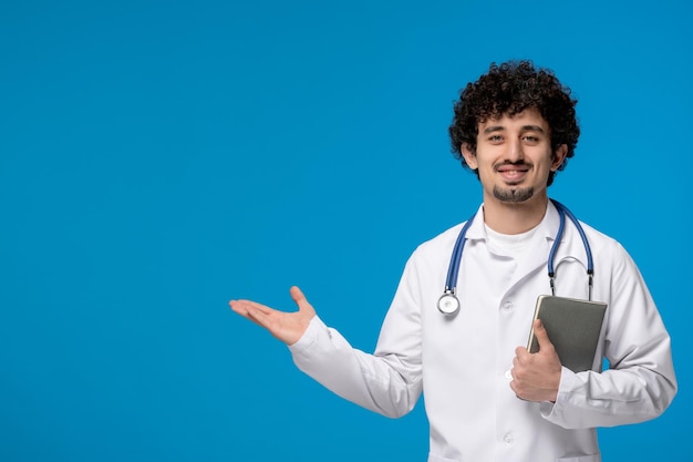 Doktersdag krullende knappe, schattige kerel in medisch uniform die een boek vasthoudt en glimlacht