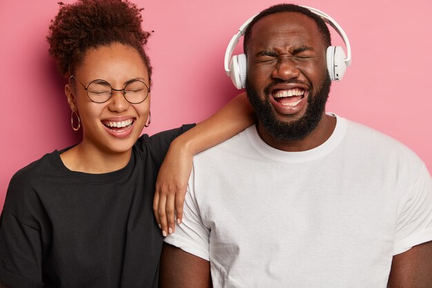 Dolblij Afro-Amerikaans stel heeft plezier, luister naar favoriete muziek in een koptelefoon, giechel over iets positiefs, draag zwart-witte kleding, geniet ervan om samen tijd door te brengen, geïsoleerd op een roze muur