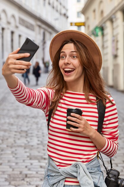 Dolblij Europese vrouwelijke reiziger in hoed, maakt selfie-portret buiten, heeft plezier tijdens excursie in de oude stad