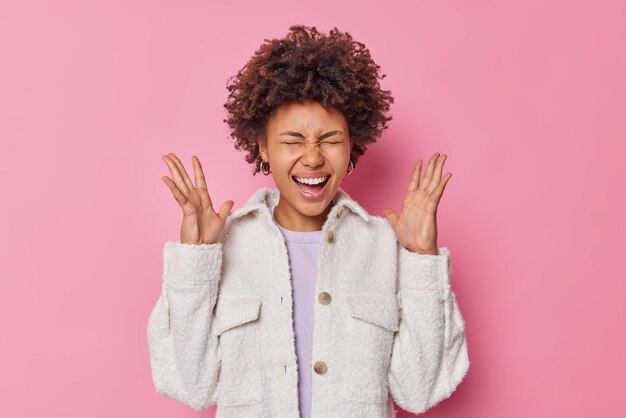 Dolblij jonge vrouw met krullend haar roept luid houdt handpalmen omhoog lacht gelukkig gekleed in bontjas blij om geweldig nieuws te horen geïsoleerd over roze muur