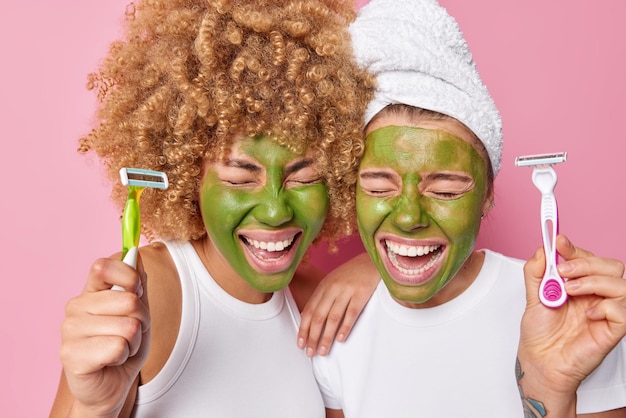 Dolblij jonge vrouwelijke modellen houden scheermesjes voor het scheren ondergaan hygiëne procedures toepassen groene natuurlijke schoonheid masker op gezicht glimlach tandjes gekleed in casual t-shirt geïsoleerd over roze achtergrond