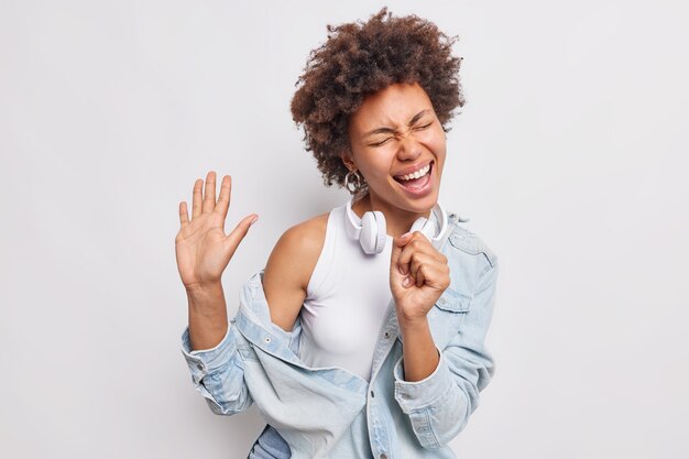 Dolblij krullende Afro-Amerikaanse vrouw houdt handpalm omhoog zingt lied houdt hand in de buurt van mond alsof microfoon denim overhemd hoofdtelefoon om nek draagt geïsoleerd over witte muur