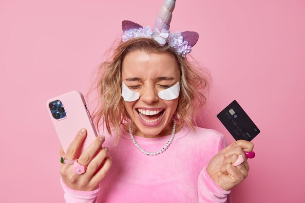 Dolblij positieve vrouw ondergaat schoonheidsprocedures past witte vlekken onder ogen toe houdt mobiele telefoon en creditcard maakt online betaling geïsoleerd over roze achtergrond Bank- en betaalconcept