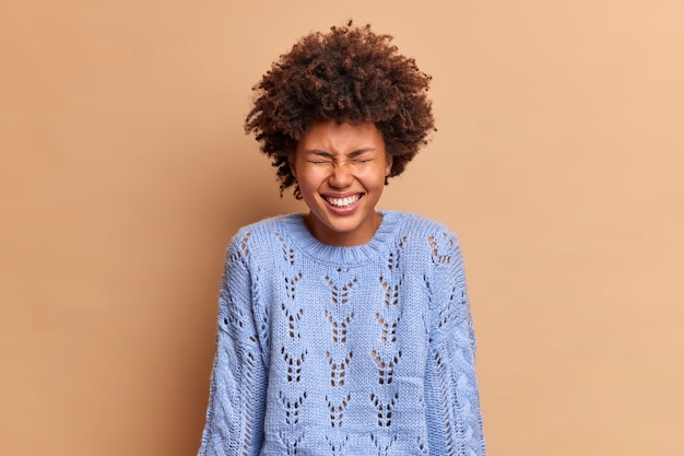 Dolblij vrouw met krullend haar lacht om iets positiefs grinnikt en sluit ogen heeft witte tanden draagt casual gebreide trui geïsoleerd over bruine muur