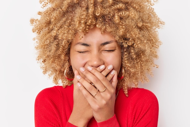 Dolblije oprechte vrouw met krullend haar lacht vreugdevol bedekt mond met handen probeert emoties te verbergen draagt casual rode trui houdt ogen gesloten poses tegen witte achtergrond Gelukkig vrouwelijk model