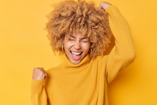 Dolblije vrouw met krullend haar maakt ja gebaar voelt opgewonden en triomfantelijk roept van vreugde draagt casual trui poses over gele studio achtergrond viert succes Ja ik heb mijn doel bereikt