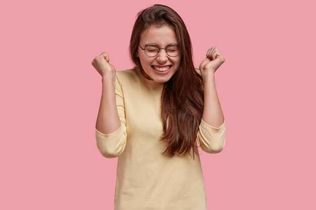 Dolblije vrouw steekt gejuich met gebalde vuisten op, geniet van succes en triomf, draagt een bril en casual outfit, modellen boven roze ruimte