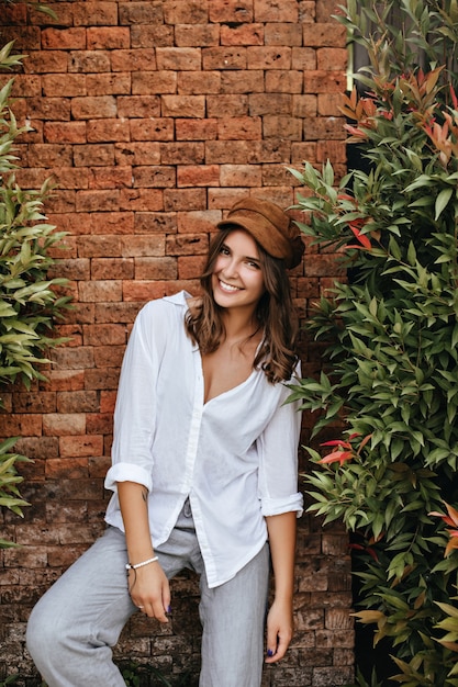 Donker blonde vrouw in oversized blouse en broek in uitstekende bui poseren naast oude bakstenen muur en bomen.