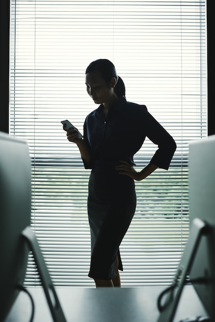 Donker silhouet van vrouw met smartphone die zich in bureau bij venster met zonneblinden bevindt