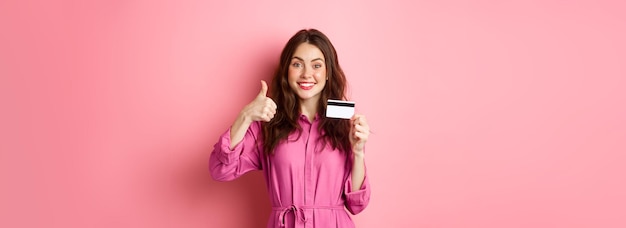 Gratis foto donkerbruin meisje dat duimen opsteekt en glimlacht en een bank aanbeveelt met een plastic creditcard die tegen een roze achtergrond staat