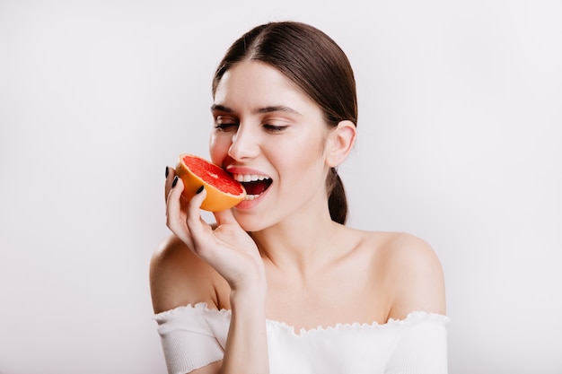 Donkerbruin meisje met een gezonde huid bijt sappige rode grapefruit. Close-upportret van vrouw in goed humeur op geïsoleerde muur.