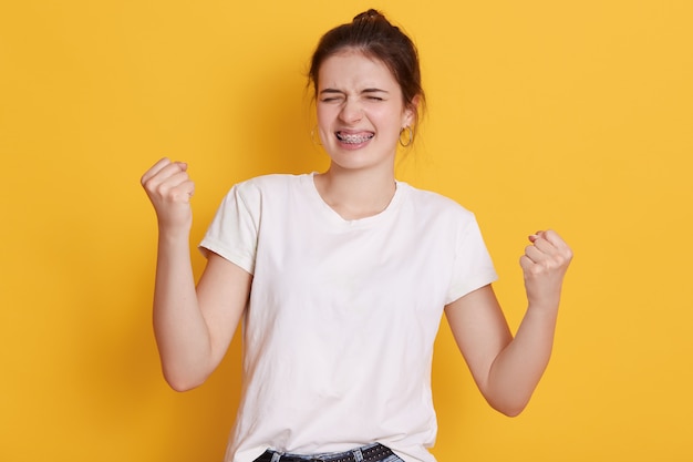 Donkerbruine jonge aantrekkelijke jonge vrouw die vuisten dichtklemmen en glimlachen, die haar succes vieren