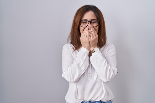 Donkerbruine vrouw die over een witte geïsoleerde achtergrond staat lachend en beschaamd giechelend mond bedekkend met handen roddels en schandaalconcept